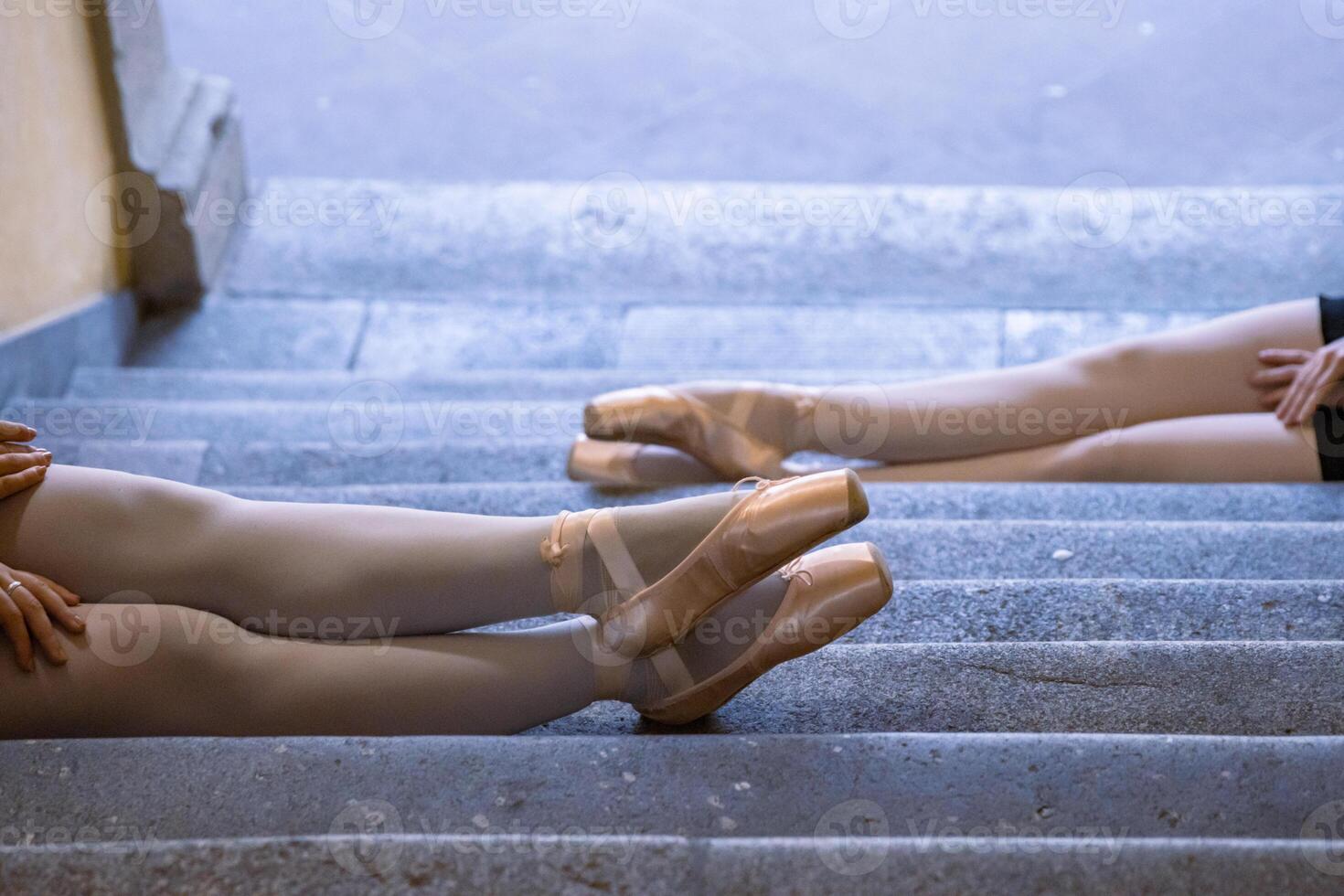 Two young ballerinas are resting on the stairs with their legs stretched on the stone steps with pointe shoes on. Ballet students are taking a brake sitting on the floor, top view. Selective focus photo