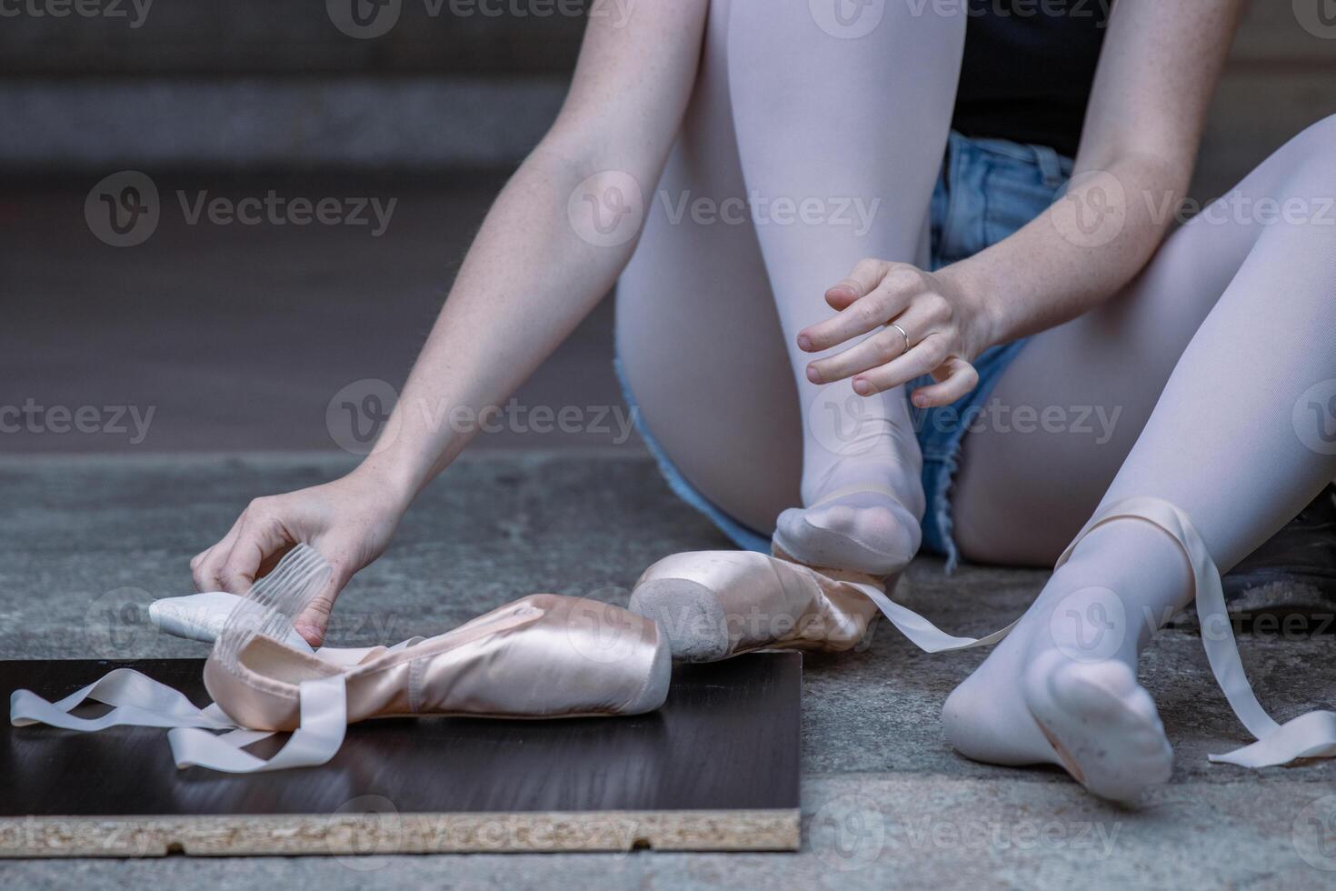 Ballerina is putting on her pointe shoes sitting on the stone floor. Ballet dancer tying ballet shoes. Dance student is preparing to the performance. Close-up. Selective focus. photo