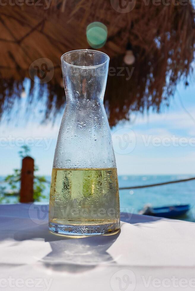Fresh fruit water in a glass pitcher on the table in the restaurant at the sea shore. Ice cold lemonade at the beach. Detail of the bottle. Drops of water. Blue sky and sea on the background. Close-up photo