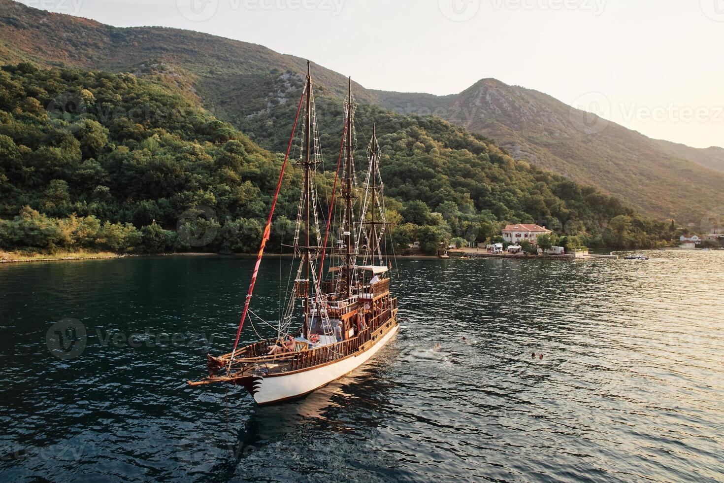 Tourists swim in the sea near a sailing yacht opposite the mountainous coast. Drone photo