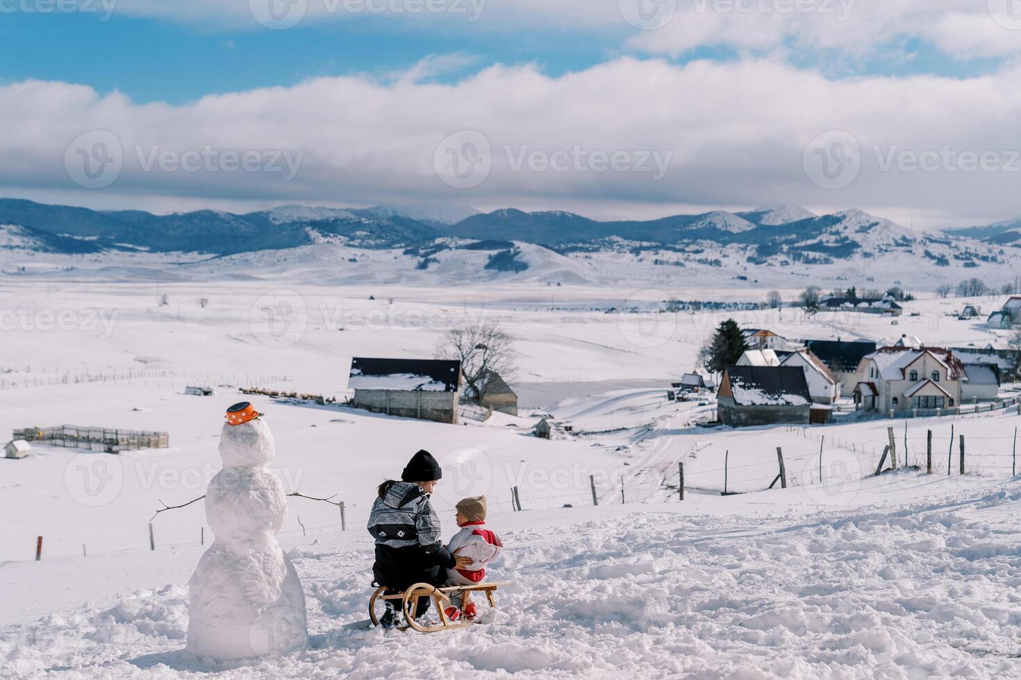Mom and little girl are sitting on a sled on a snow-capped plain near a snowman and look at each other. Back view photo