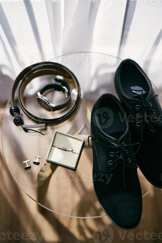 Wedding rings in a box lie on a glass table next to the groom accessories. Top view photo