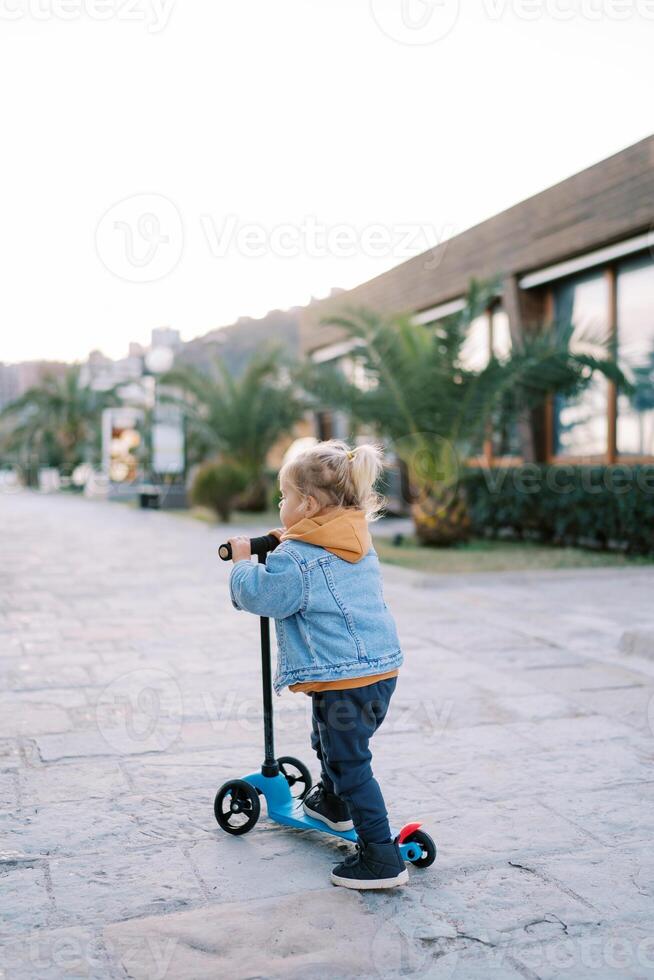 Little girl rides a scooter on the road past houses. Back view photo