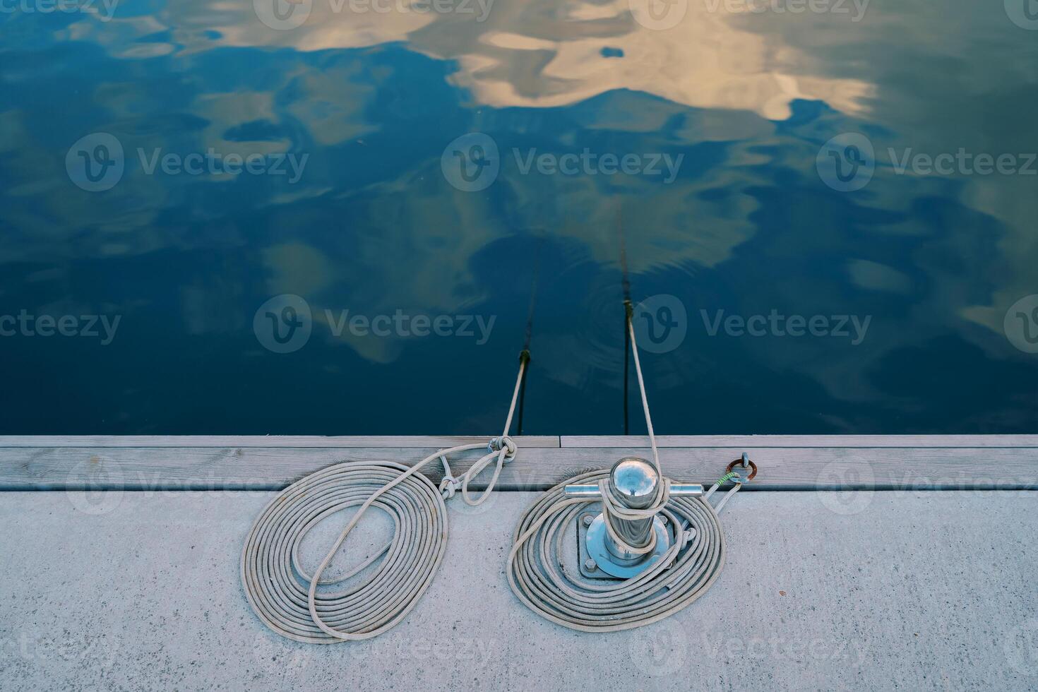 Mooring ropes are wrapped around a metallic shiny bollard on a pier by the sea photo