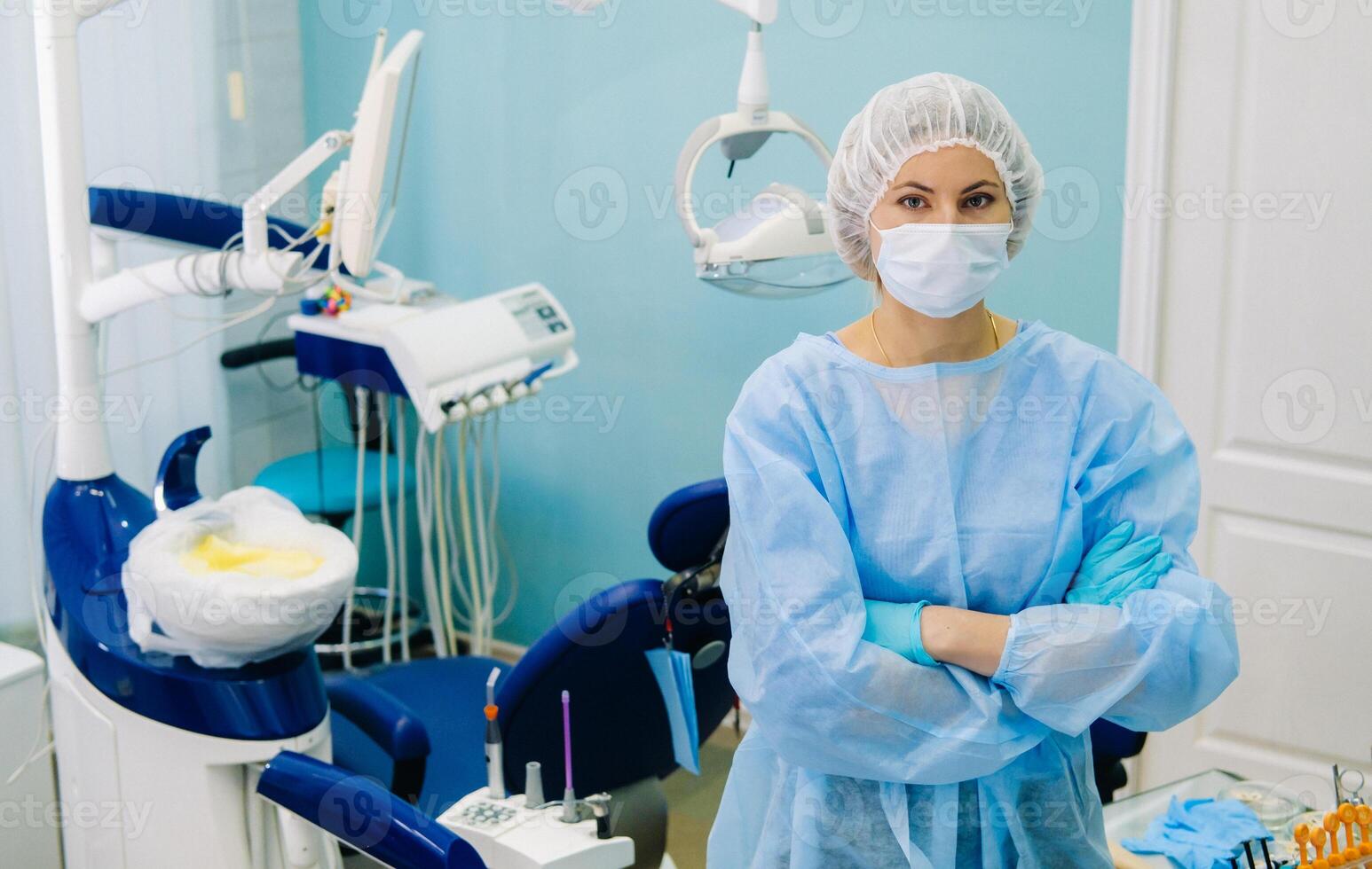 un hembra dentista vistiendo un médico máscara y caucho guantes poses para el cámara y pliegues su brazos en su oficina foto