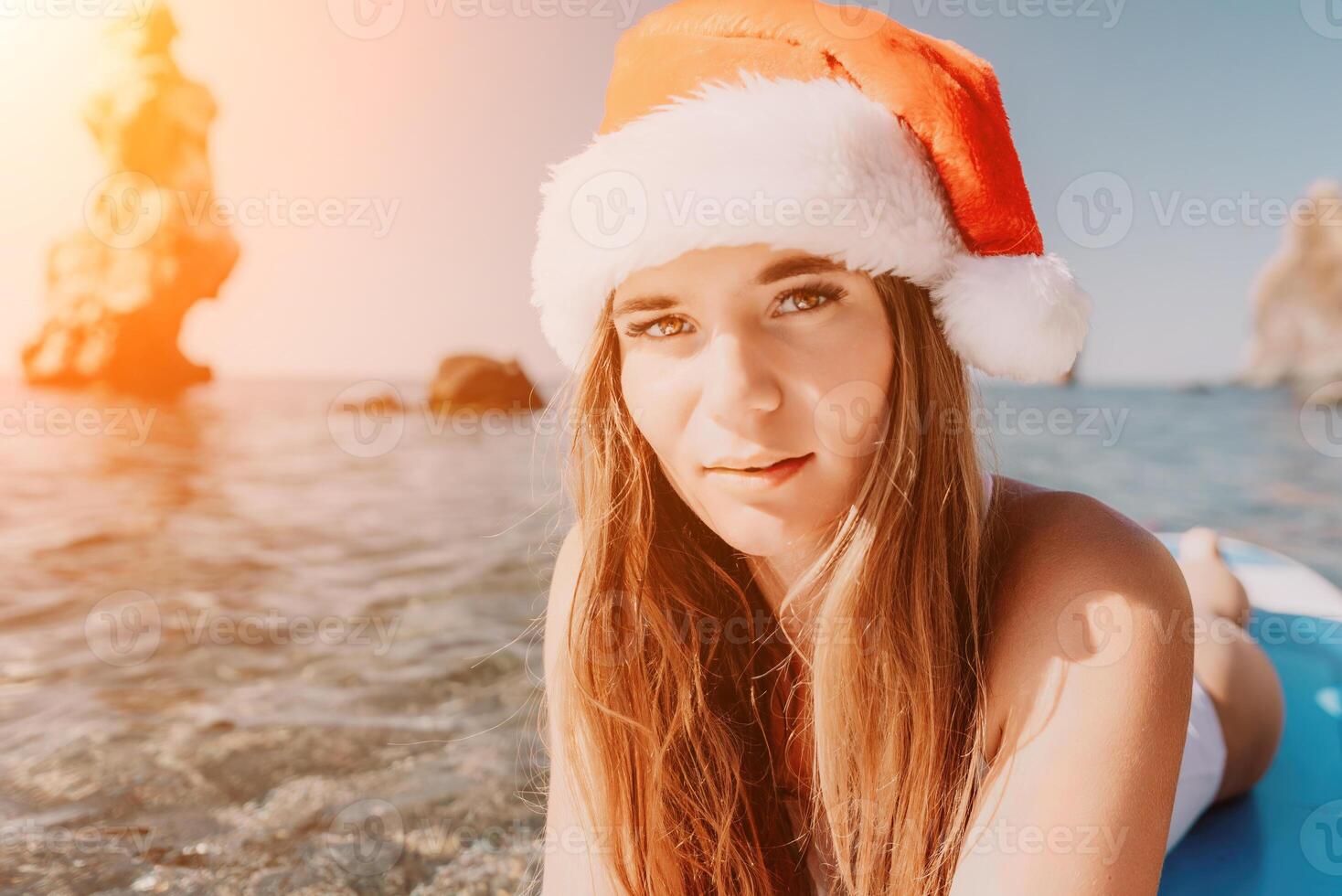 mujer mar sorber. cerca arriba retrato de contento joven caucásico mujer con largo pelo en Papa Noel sombrero mirando a cámara y sonriente. linda mujer retrato en un blanco bikini posando en cenar tablero en el mar foto