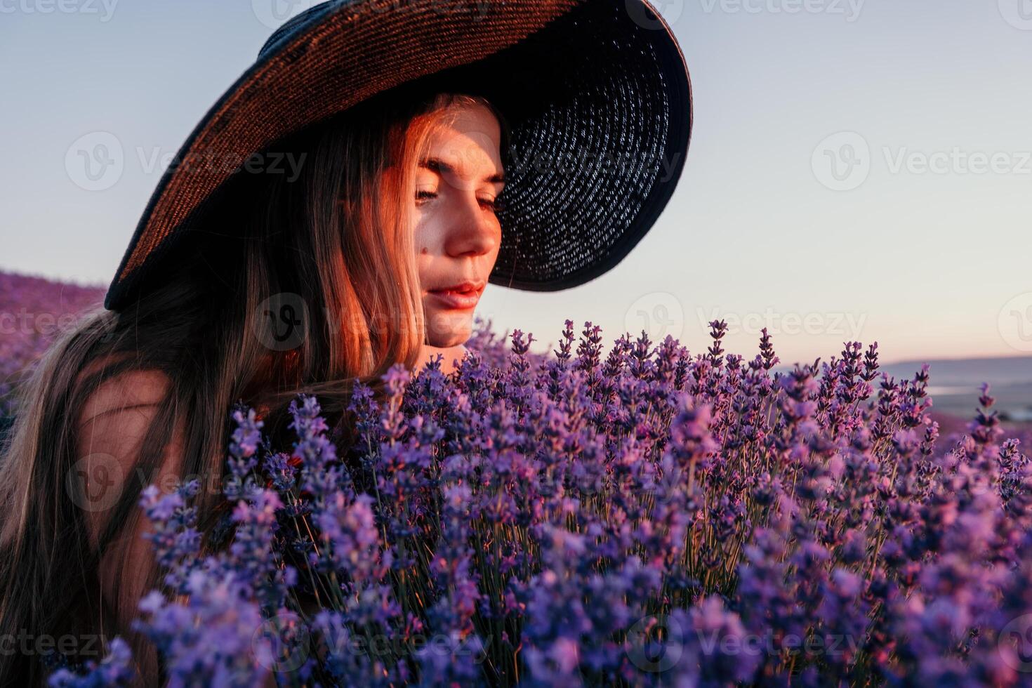 Woman lavender field. Happy carefree woman in black dress and hat with large brim smelling a blooming lavender on sunset. Perfect for inspirational and warm concepts in travel and wanderlust. Close up photo