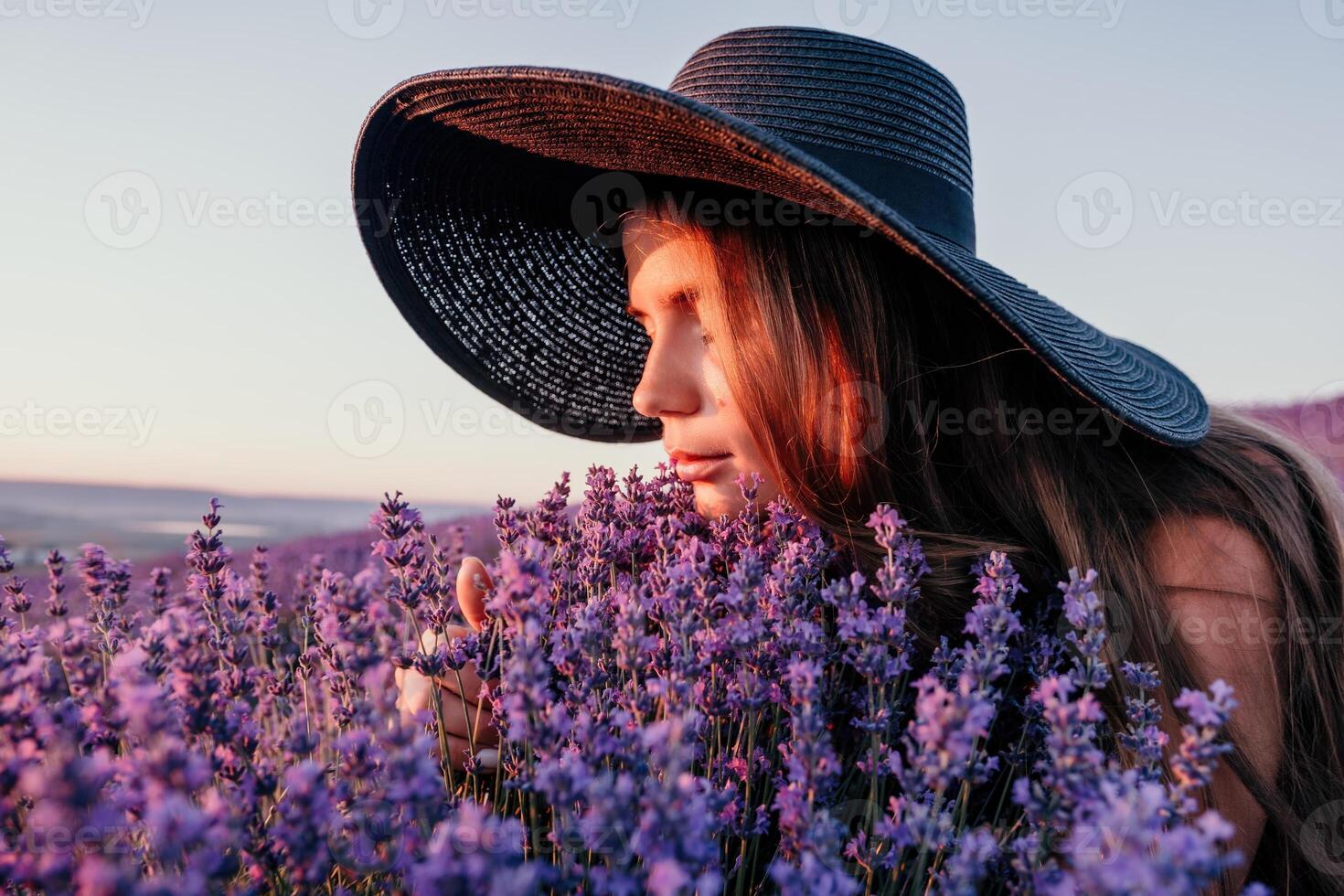 Woman lavender field. Happy carefree woman in black dress and hat with large brim smelling a blooming lavender on sunset. Perfect for inspirational and warm concepts in travel and wanderlust. Close up photo