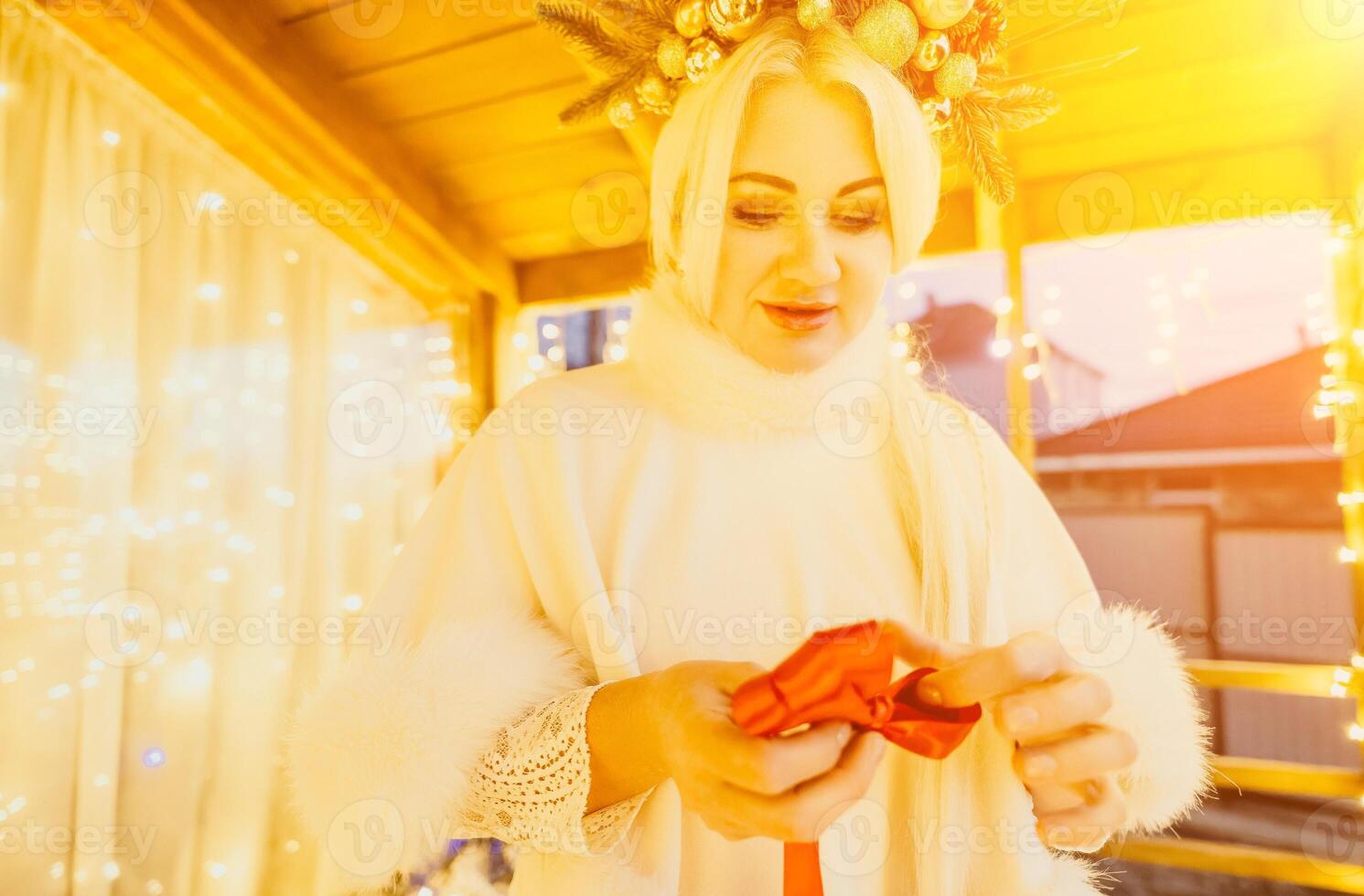 A woman in a white dress is holding a gold box with a red ribbon. She is wearing a crown on her head. The woman appears to be opening the gift box. photo