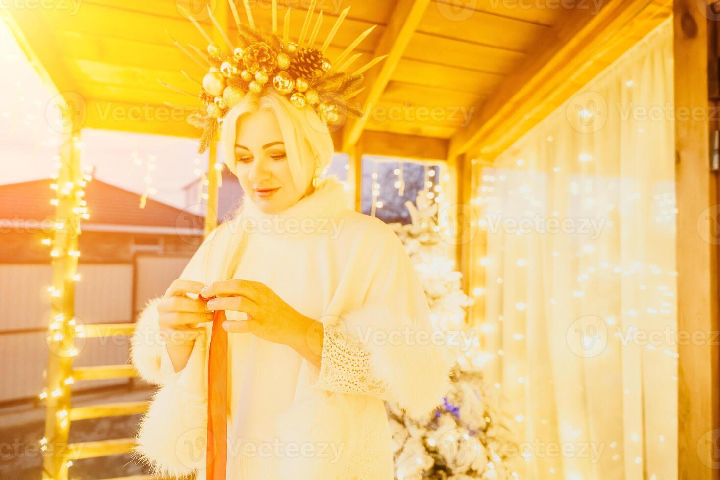 A woman in a white dress is holding a gold box with a red ribbon. She is wearing a crown on her head. The woman appears to be opening the gift box. photo