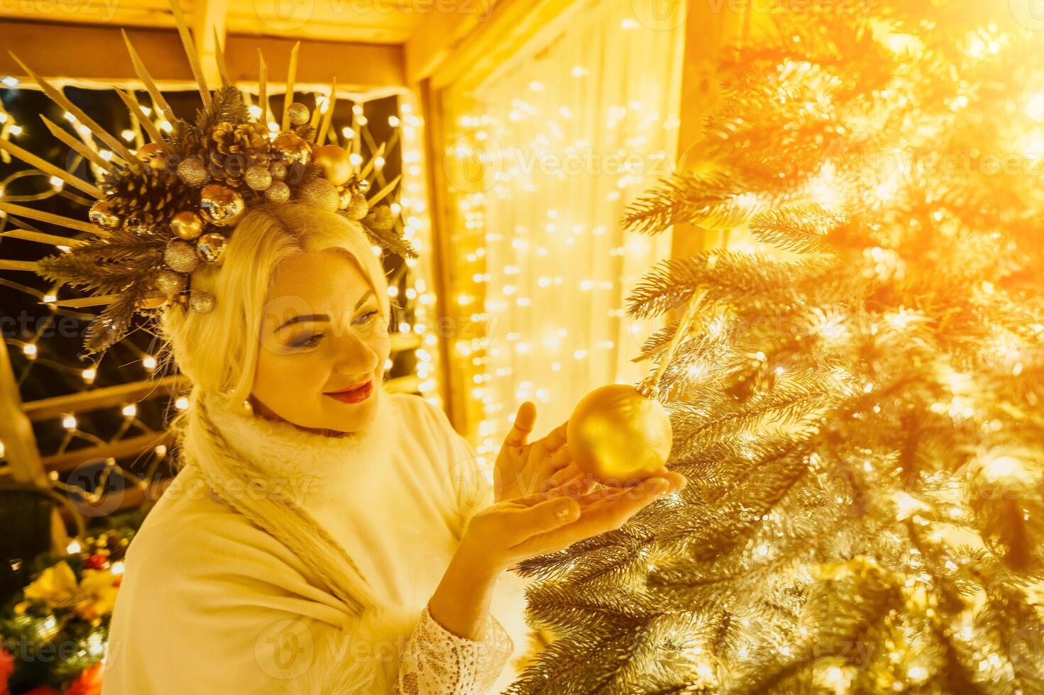 A blonde woman in white dress and a crown of gold ornaments decorate Christmas tree with gold ornaments and lights. The tree is decorated with gold balls and is lit up with lights. photo