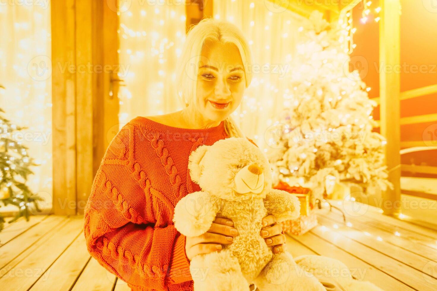 Happy smiling Woman in a red sweater sits on a festive porch, holding a teddy bear next to Snow-covered Christmas tree with bright white lights, exuding contented enjoyment in a cozy, cheerful smile photo