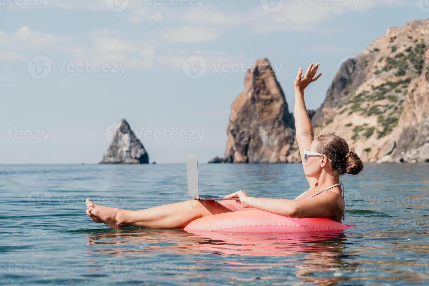 Woman freelancer works on laptop swimming in sea on pink inflatable ring. Happy tourist in sunglasses floating on inflatable donut and working on laptop computer in calm ocean. Remote working anywhere photo