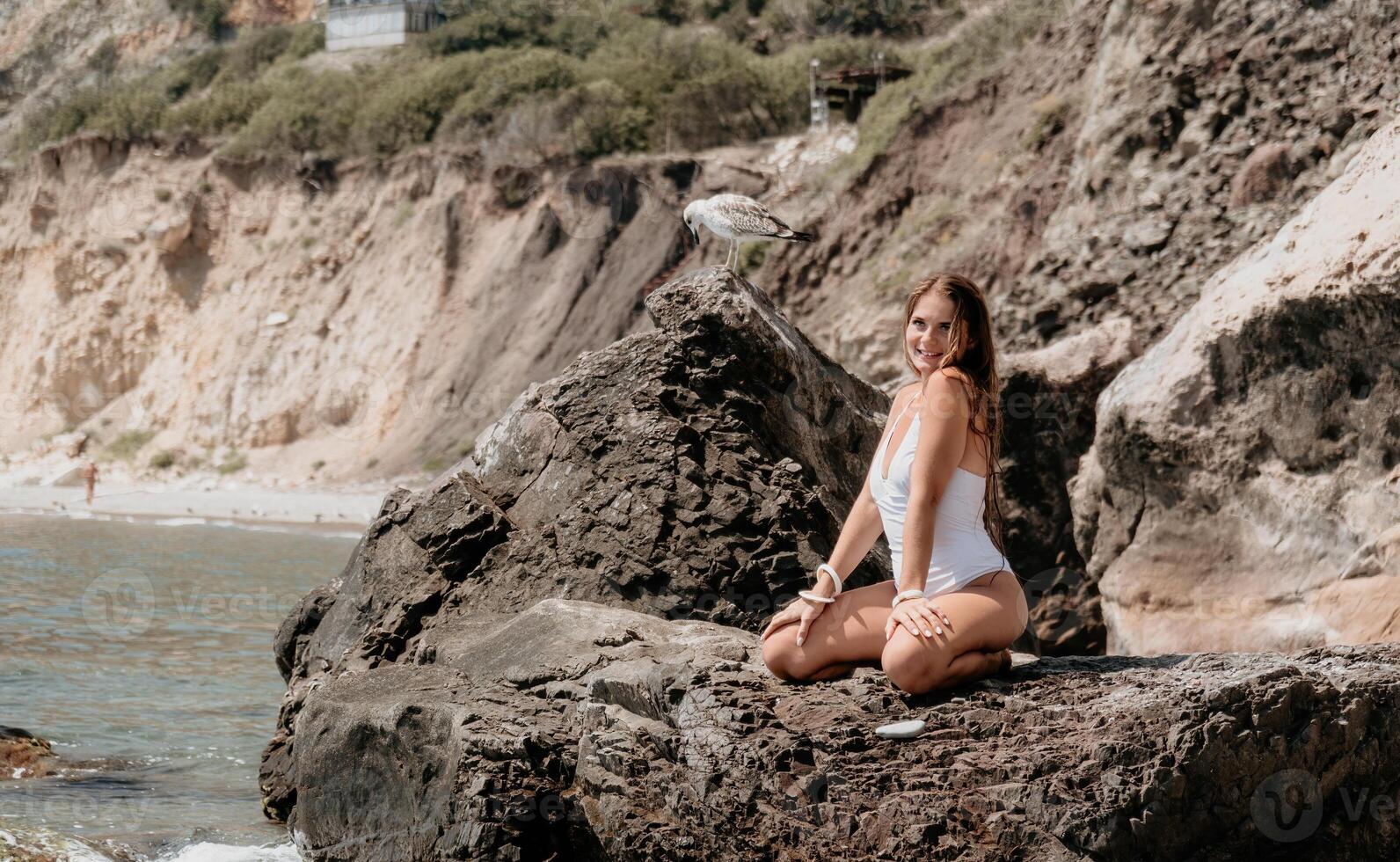 mujer viaje mar. contento turista en blanco bikini disfrutar tomando imagen al aire libre para recuerdos. mujer viajero posando en el playa a mar rodeado por volcánico montañas, compartiendo viaje aventuras viaje foto