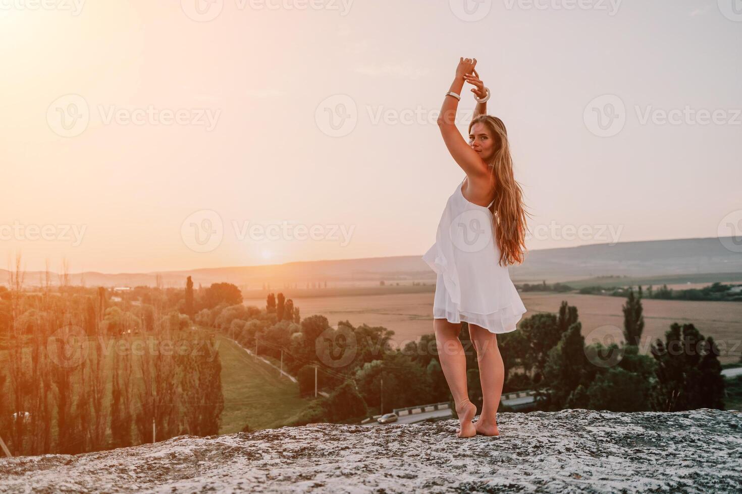 Happy woman standing with her back on the sunset in nature in summer with open hands. Romantic beautiful bride in white boho dress posing with mountains on sunset photo