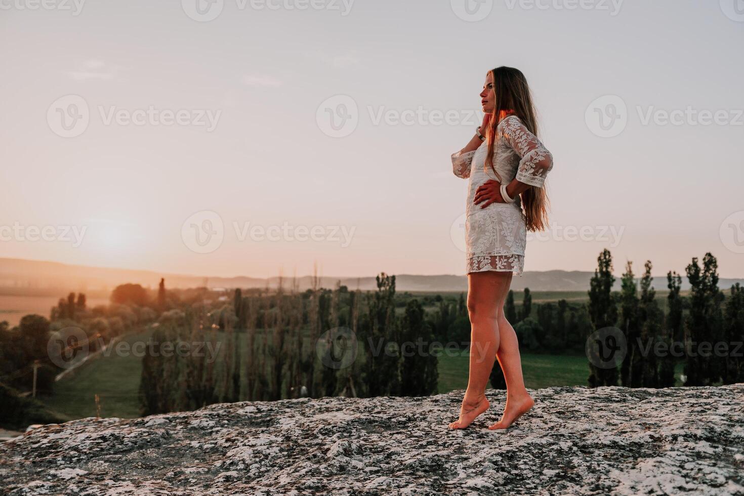 Happy woman in white boho dress on sunset in mountains. Romantic woman with long hair standing with her back on the sunset in nature in summer with open hands. Silhouette. Nature. Sunset. photo