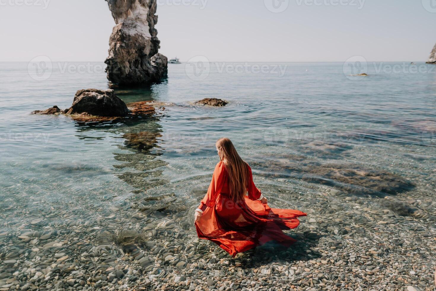 Woman travel sea. Happy tourist in red dress enjoy taking picture outdoors for memories. Woman traveler posing on the rock at sea bay surrounded by volcanic mountains, sharing travel adventure journey photo