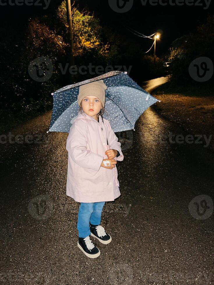 Little girl in a coat with an umbrella over her head stands on wet asphalt near a luminous lantern photo