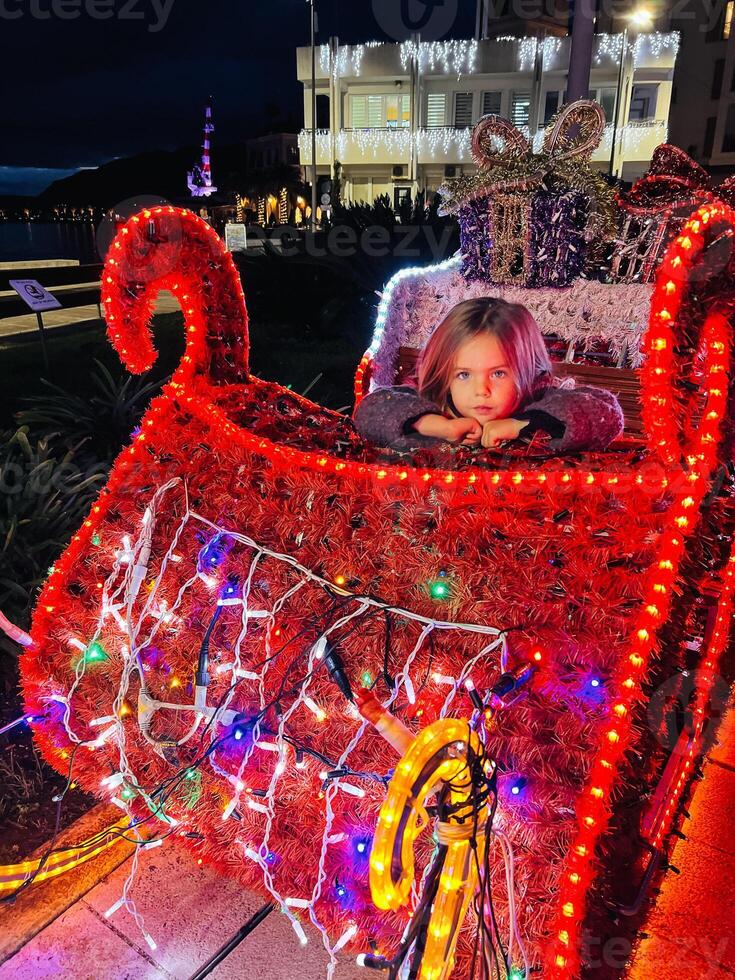 Little girl sits in Santa sleigh, with luminous garlands photo