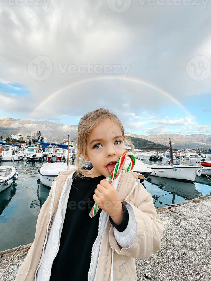 pequeño niña lame un pirulí en un muelle en contra el fondo de un arco iris foto