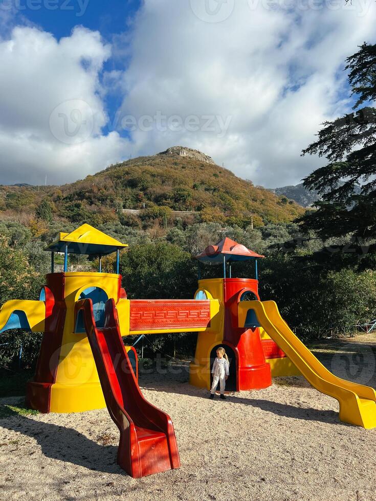 pequeño niña soportes cerca un vistoso patio de recreo con diapositivas y torres foto