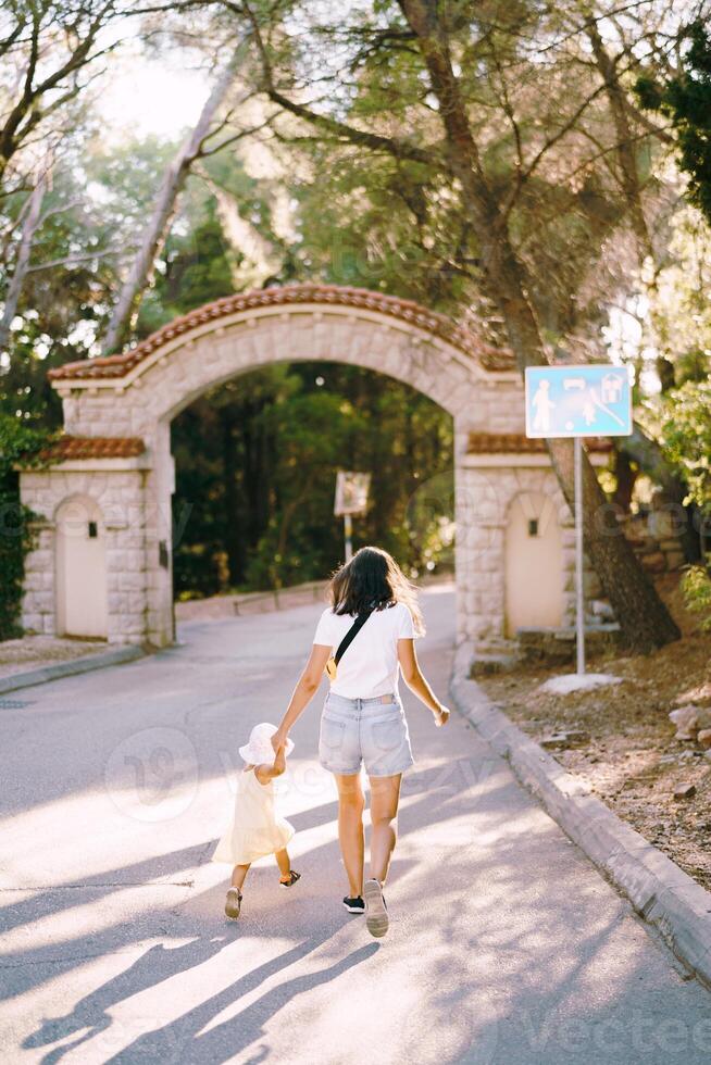 madre con un pequeño hija correr a lo largo el parque la carretera a el portón participación manos. espalda ver foto