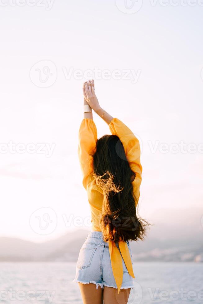 Girl in shorts and a blouse stands with her hands up by the sea. Back view photo
