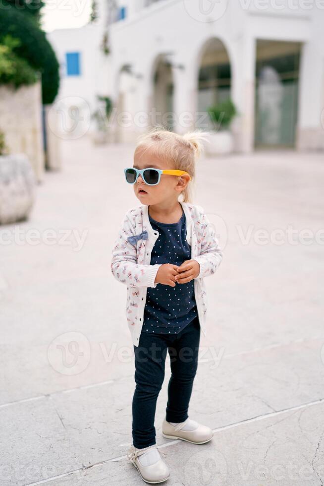 pequeño niña en Gafas de sol soportes en el yarda y mira lejos foto