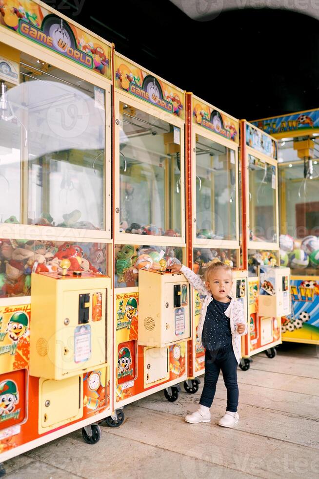 Little girl stands near a claw slot machine filled with toys photo