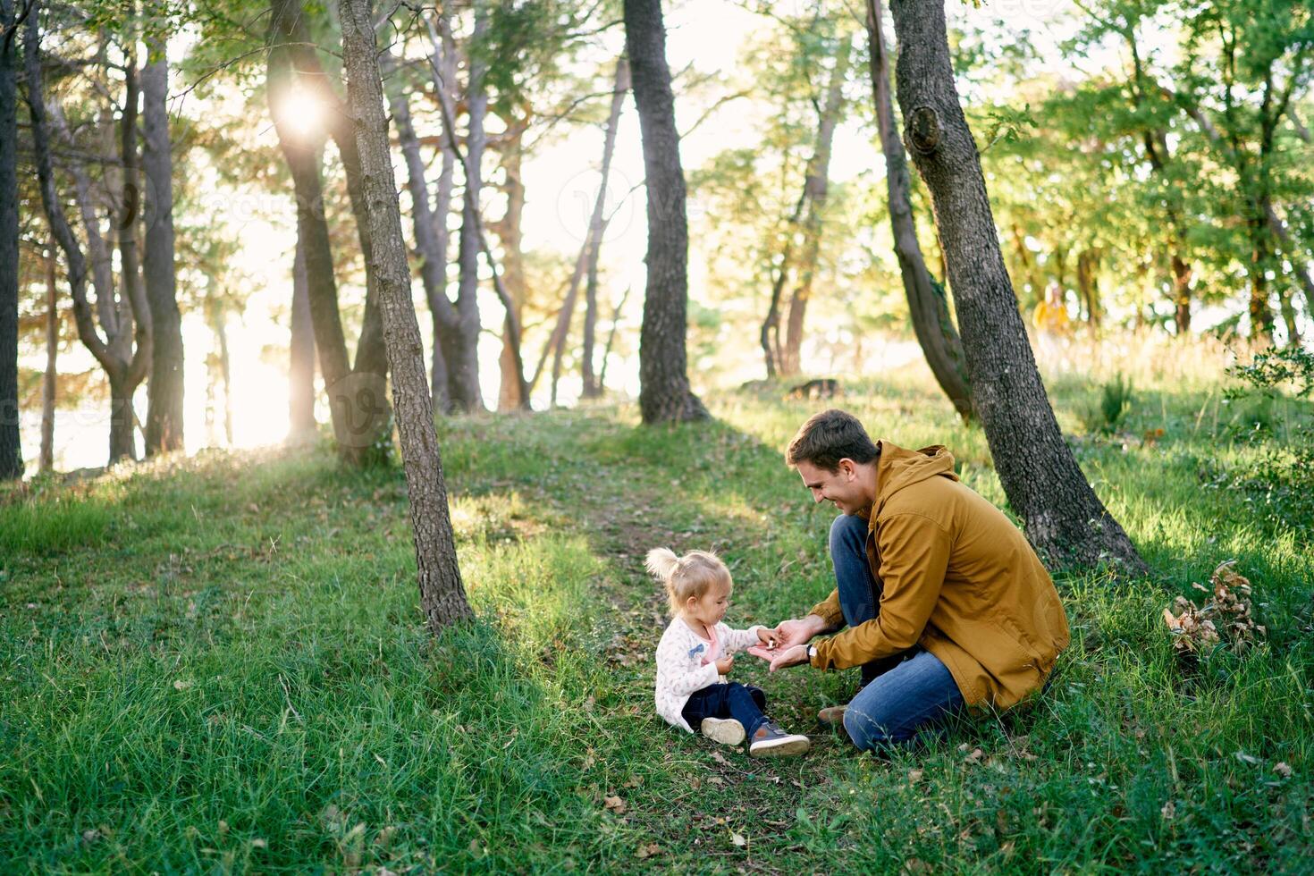 Little girl takes pebbles from the palms of her dad while sitting on a green lawn in the park photo