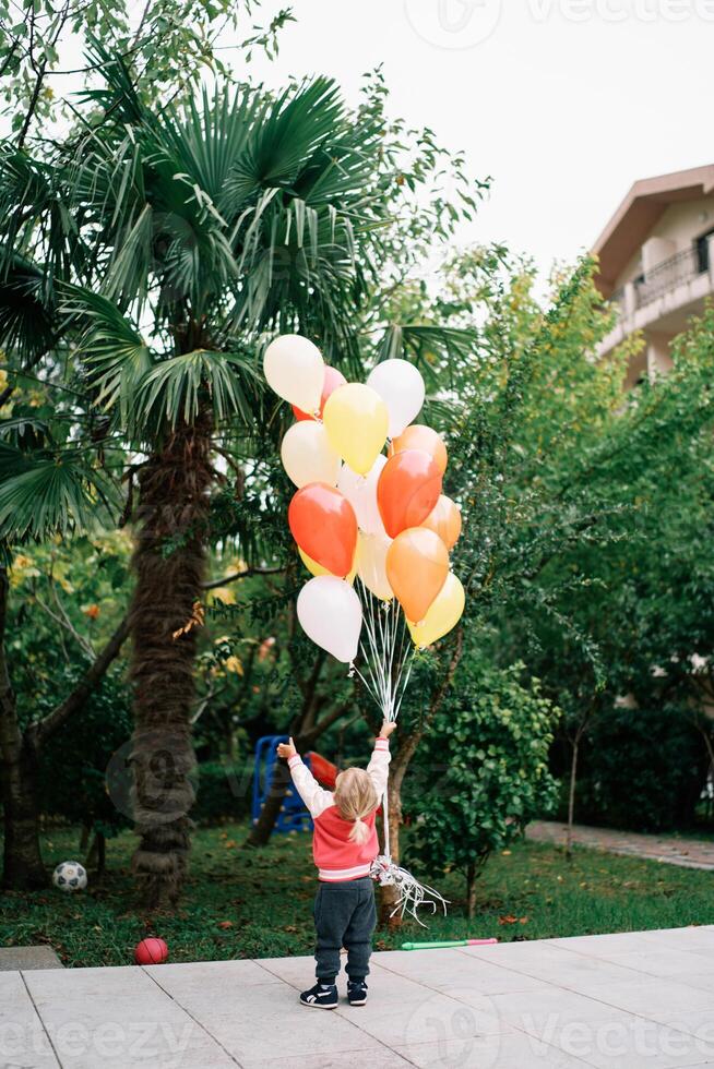 pequeño niña soportes con su mano elevado con un manojo de globos en el jardín y mira arriba. espalda ver foto