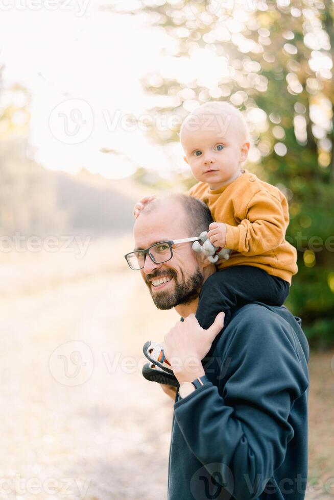 Happy dad with a little boy on his shoulders stands half-turned in a clearing photo