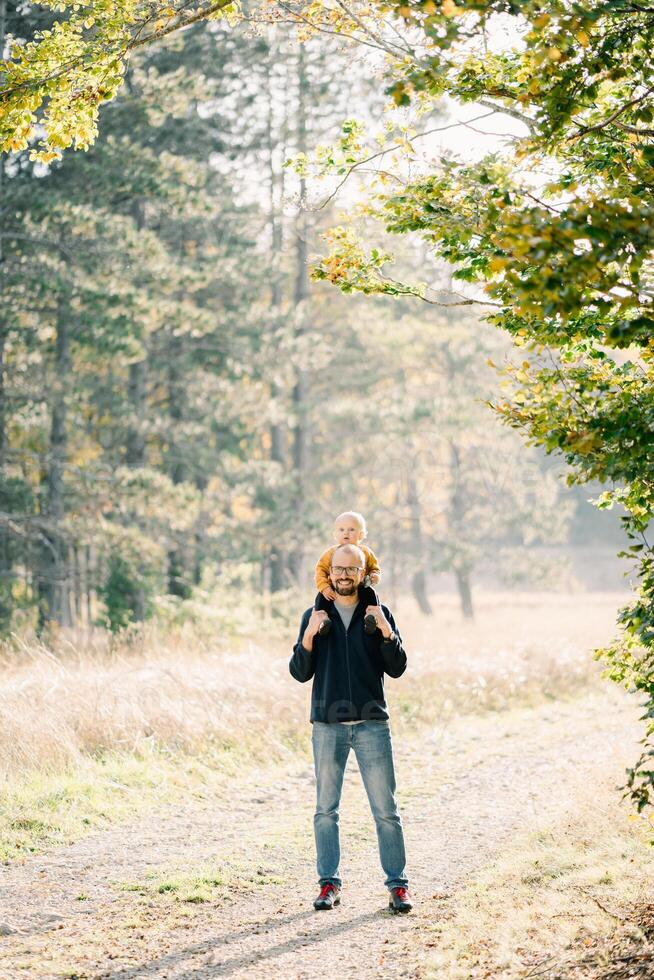 Smiling dad with a little son on his shoulders stands on a country road in a sunny forest photo