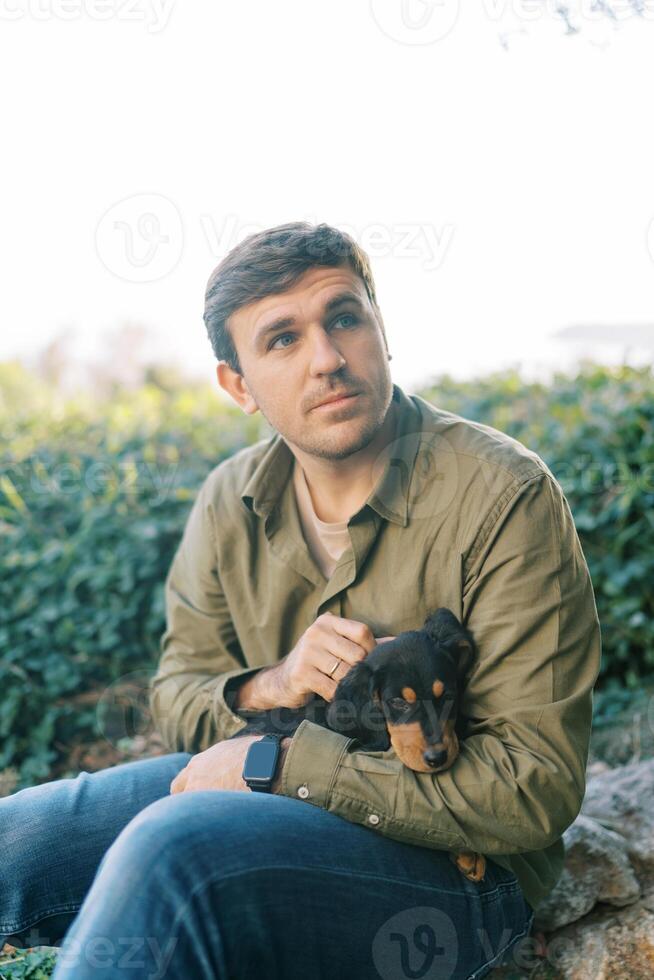 Young man sits in a park and strokes a black puppy on his knees while looking away photo