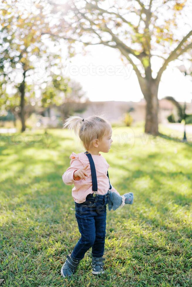 Little girl with a soft toy in her hand stands half-turned on a green lawn photo