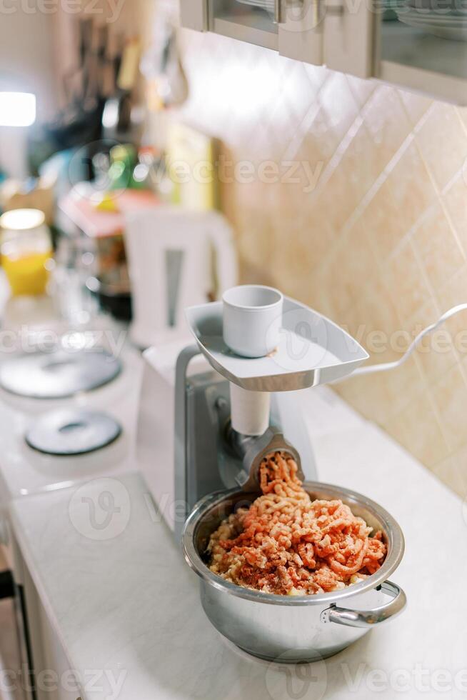 Pot with minced meat is on the table in front of the electric meat grinder photo