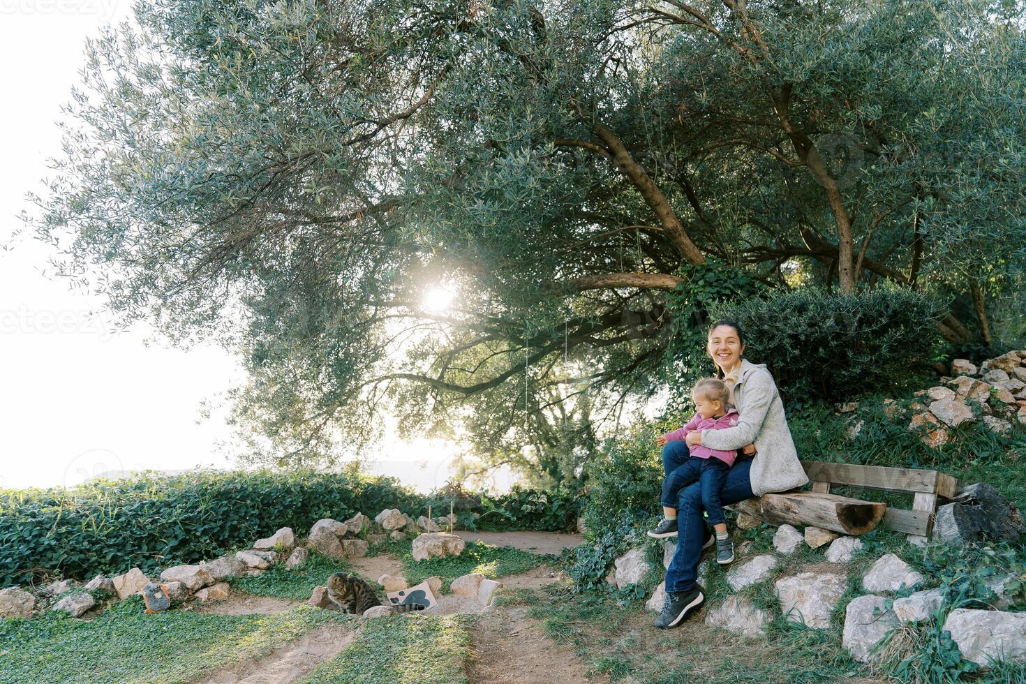 Smiling mother with a little girl in her arms sits on a park bench next to a tabby cat photo