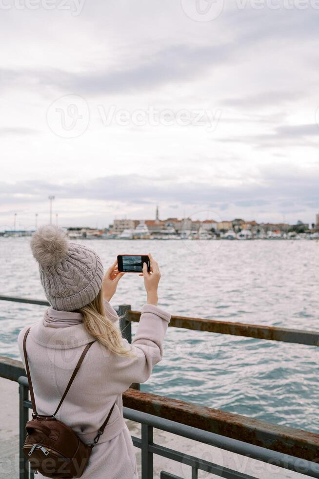 Girl with a backpack stands on the pier at the railing and takes pictures of houses on the seashore with a smartphone. Back view photo
