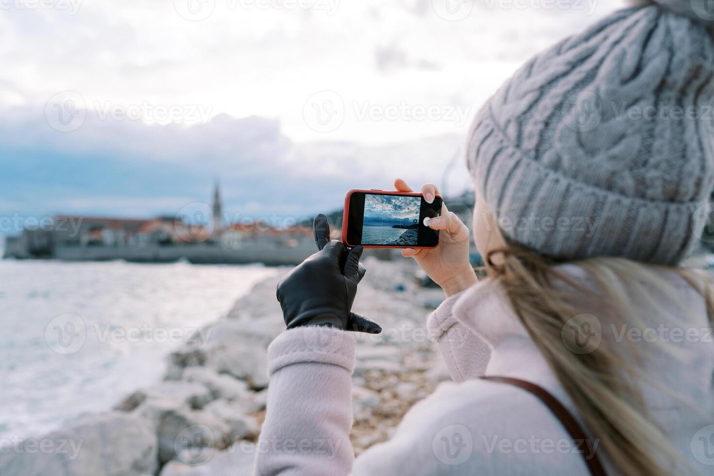 Young woman stands on the shore and shoots the sea with a smartphone. Back view photo