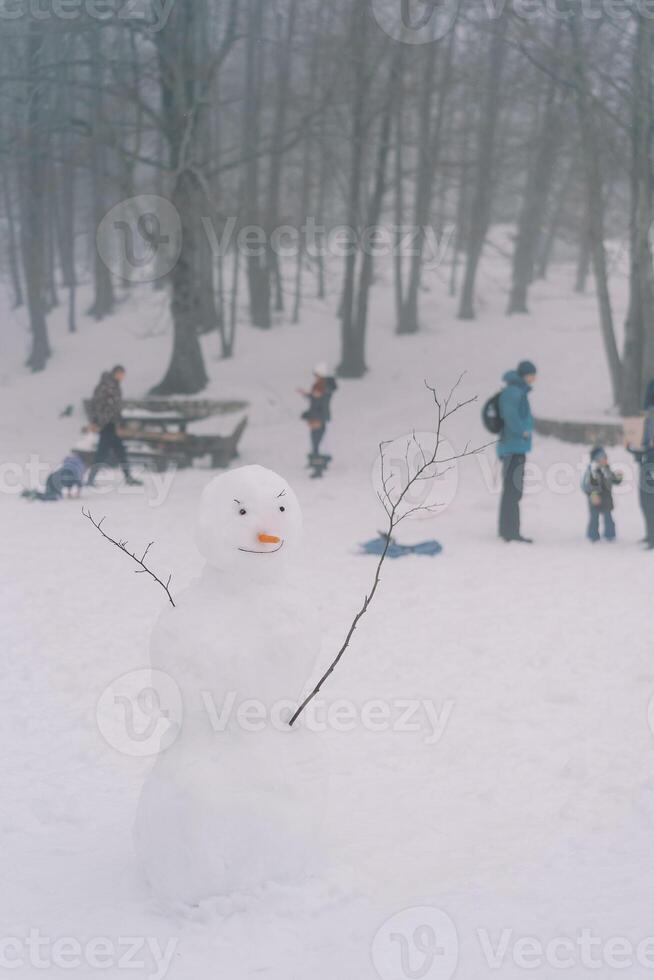 Smiling snowman with hands-twigs stands in a snowy forest against the background of people walking photo