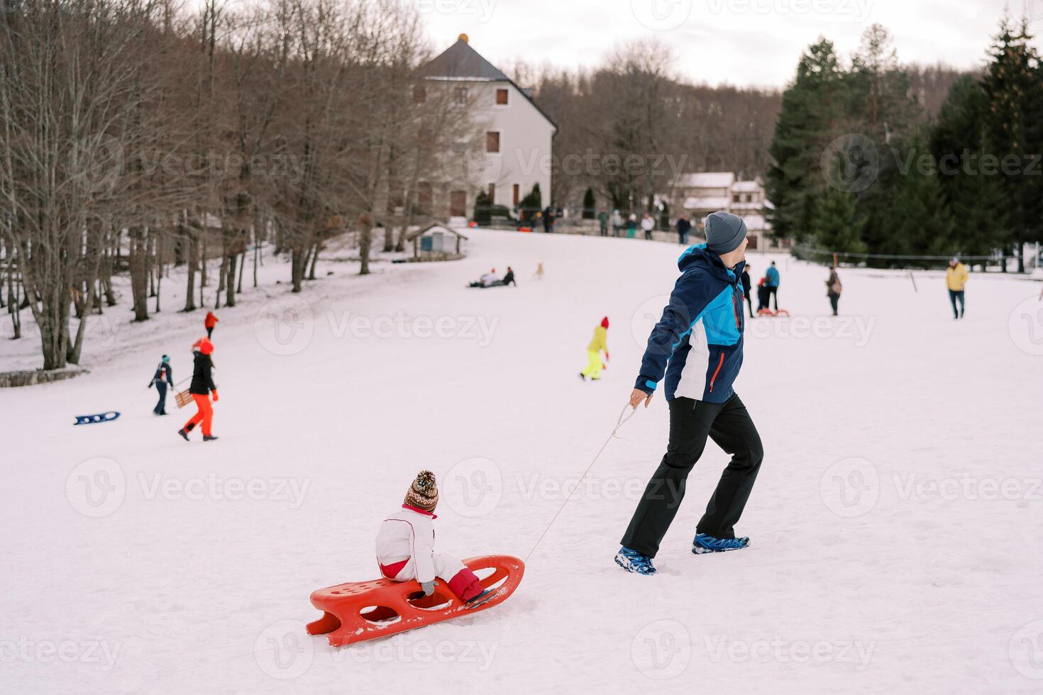 Dad drags a child in a sled by a rope up a snowy hill with resting parents with children. Back view photo