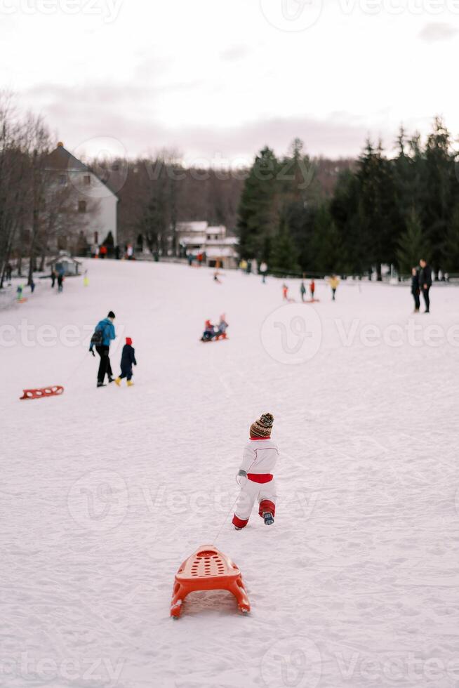 Small child in overalls drags a sled along a rope on a hill. Back view photo