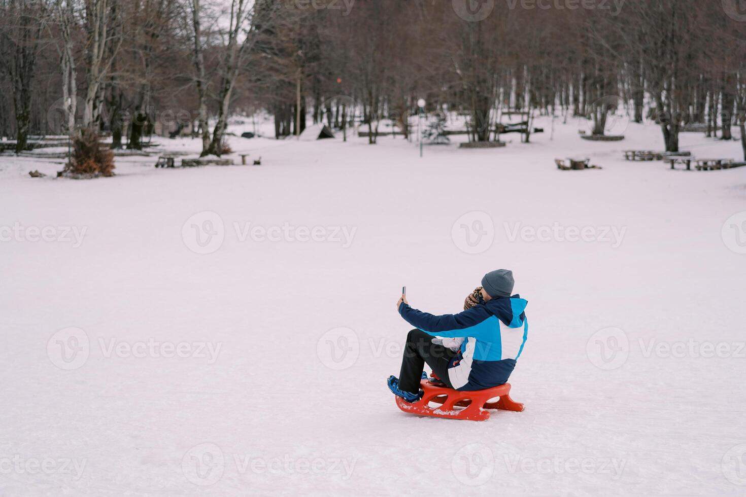 Dad taking smartphone selfie of himself and little kid sitting on sled on snowy lawn. Back view photo