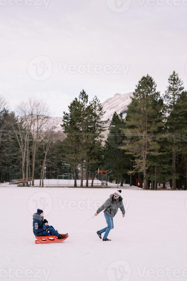 Dad is carrying mom with a child on a sled across a snowy plain looking back. Side view photo