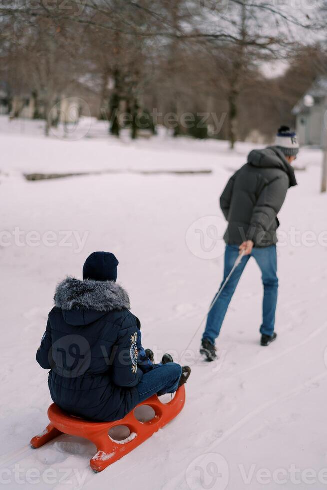Dad is pulling a sled with mom and a child on a string through the snow. Back view photo