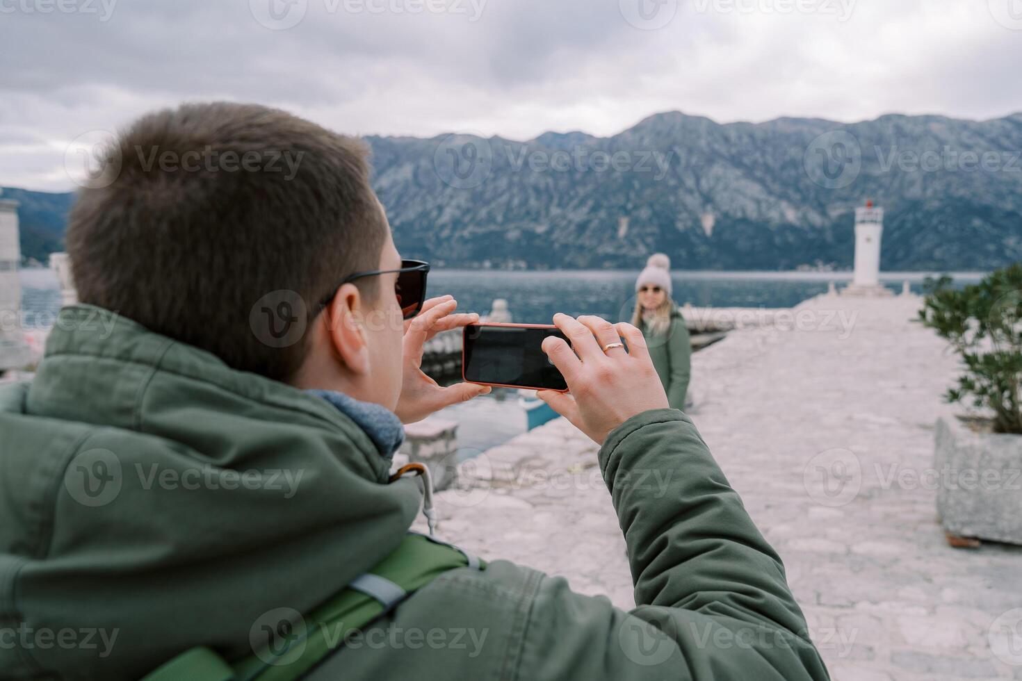 Husband takes a picture of his wife on a pier with a lighthouse in the background. Back view photo