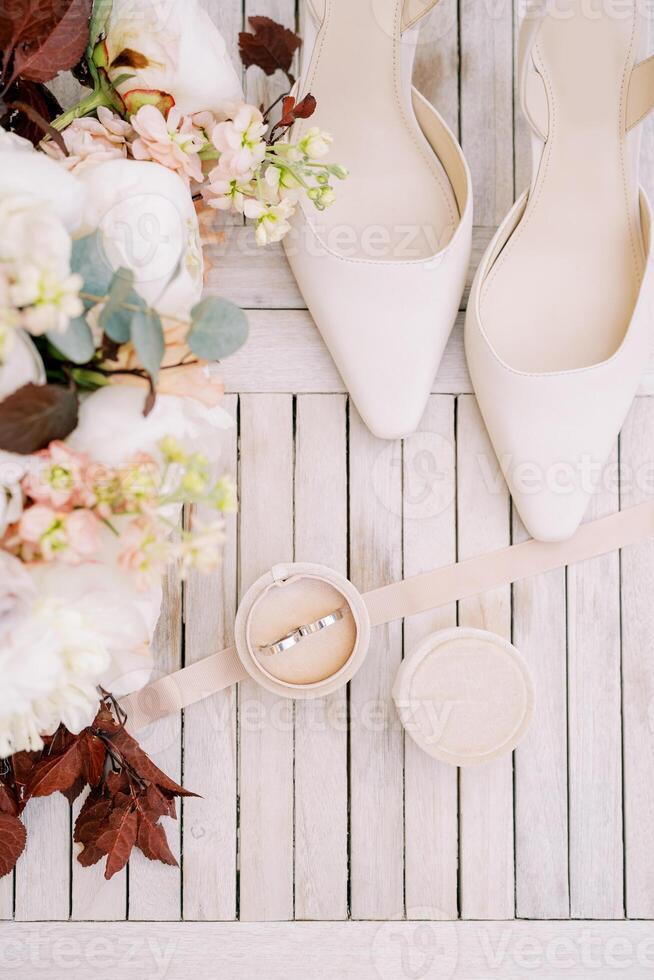 Wedding rings in a box stand on the table near the bride shoes and a bouquet of flowers. Top view photo