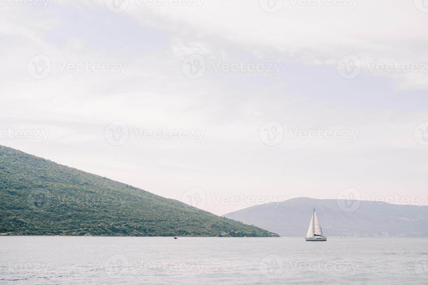 Sailing yacht sails on the sea past the green hills against the backdrop of a mountain range in a light haze photo