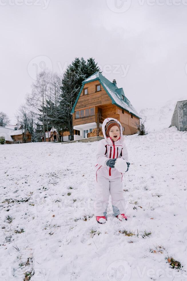 Little laughing girl with a snowball in her hands stands on a snowy slope near a wooden house photo