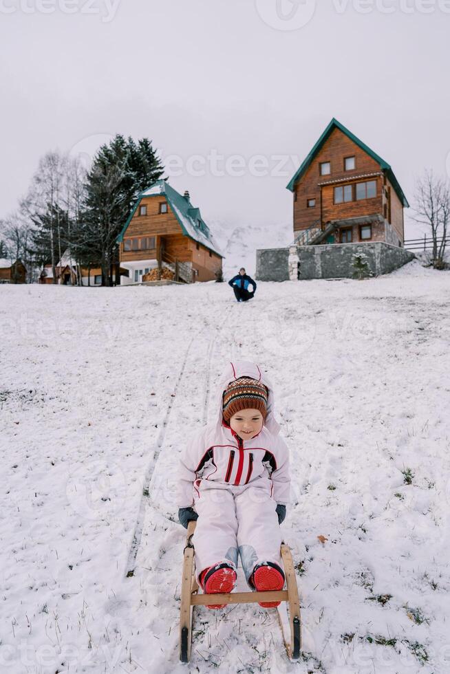 papá se sienta fuera de el casa y relojes un pequeño niña trineo abajo un Nevado colina foto